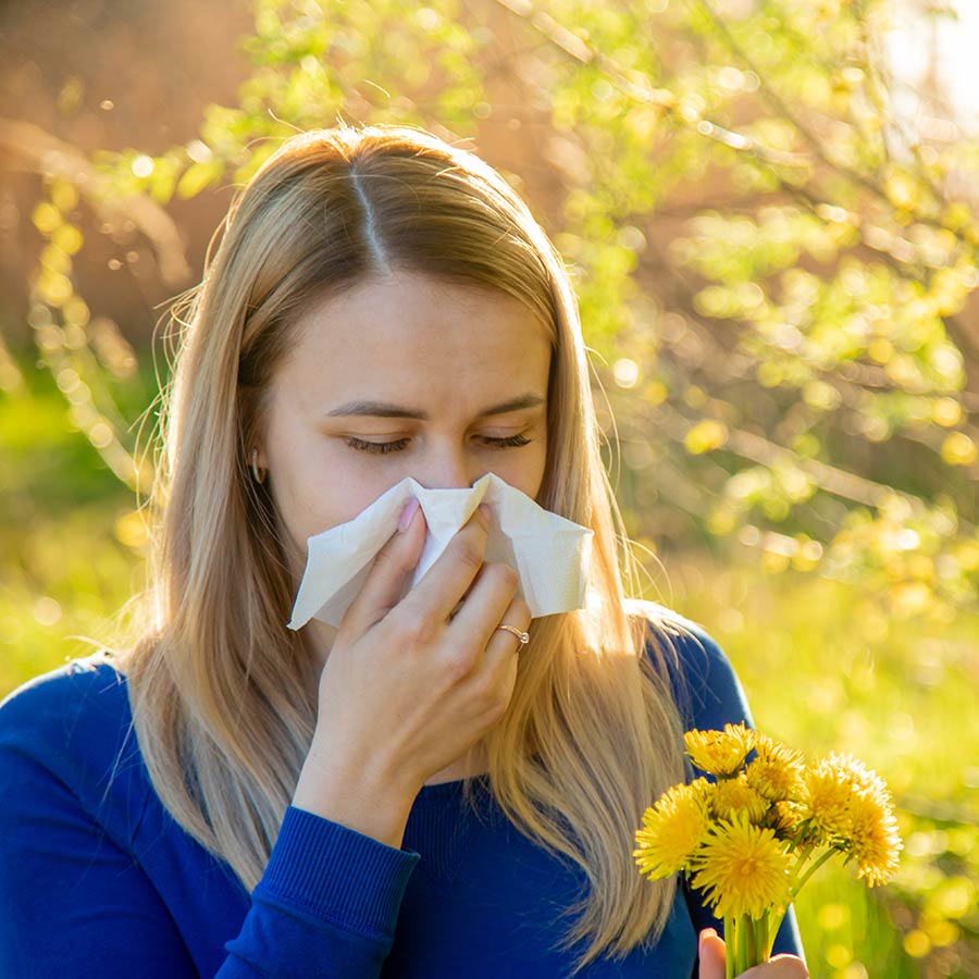 Eine junge Frau steht in Mitten von Blumen und blühenden Bäumen und niest in ein Taschentuch.